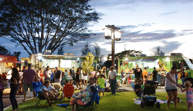 A large social gathering in front of a row of food trucks at an outdoor park at dusk.