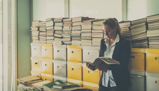 Worker reading a file stands in front of stacked file boxes.