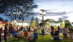 A large social gathering in front of a row of food trucks at an outdoor park at dusk.