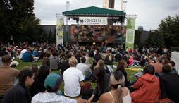 A crowd is gathered and sitting on the grass while a band plays in a covered area at the Seattle Center