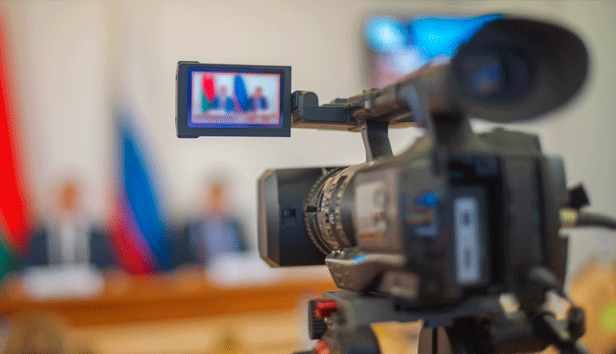 Two officials sitting at a dais during an open public meeting as seen through the lens of a video camera