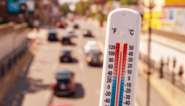 A thermometer showing a temperature of 100+ against a background of an urban street