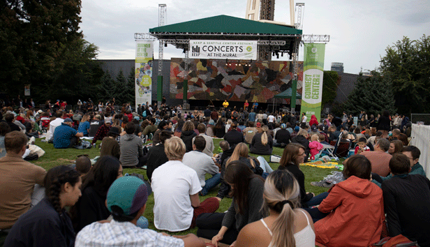 A crowd is gathered and sitting on the grass while a band plays in a covered area at the Seattle Center