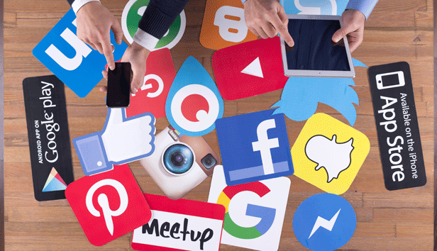 Shows a group of social media icons against a table at which two people are working; one on a cell phone and another on an iPad.