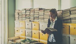 Worker reading a file stands in front of stacked file boxes.