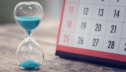 An hourglass filled with sand sits in front of a calendar