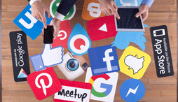 Shows a group of social media icons against a table at which two people are working; one on a cell phone and another on an iPad.